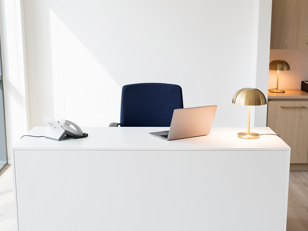 The office reception desk with a laptop, a desk phone and a small brass lamp, navy chair empty, soft daylight from the w